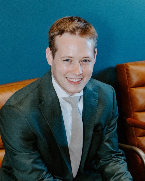 Colby Taylor in a dark suit, smiling, seated in front of a teal wall